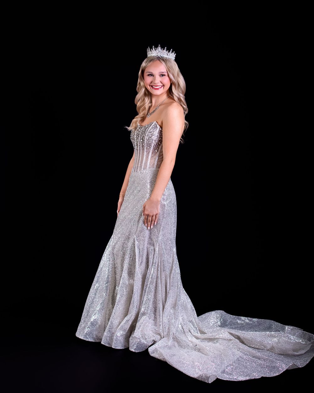 Young woman in sparkling silver mermaid gown and crystal crown poses elegantly against black background.