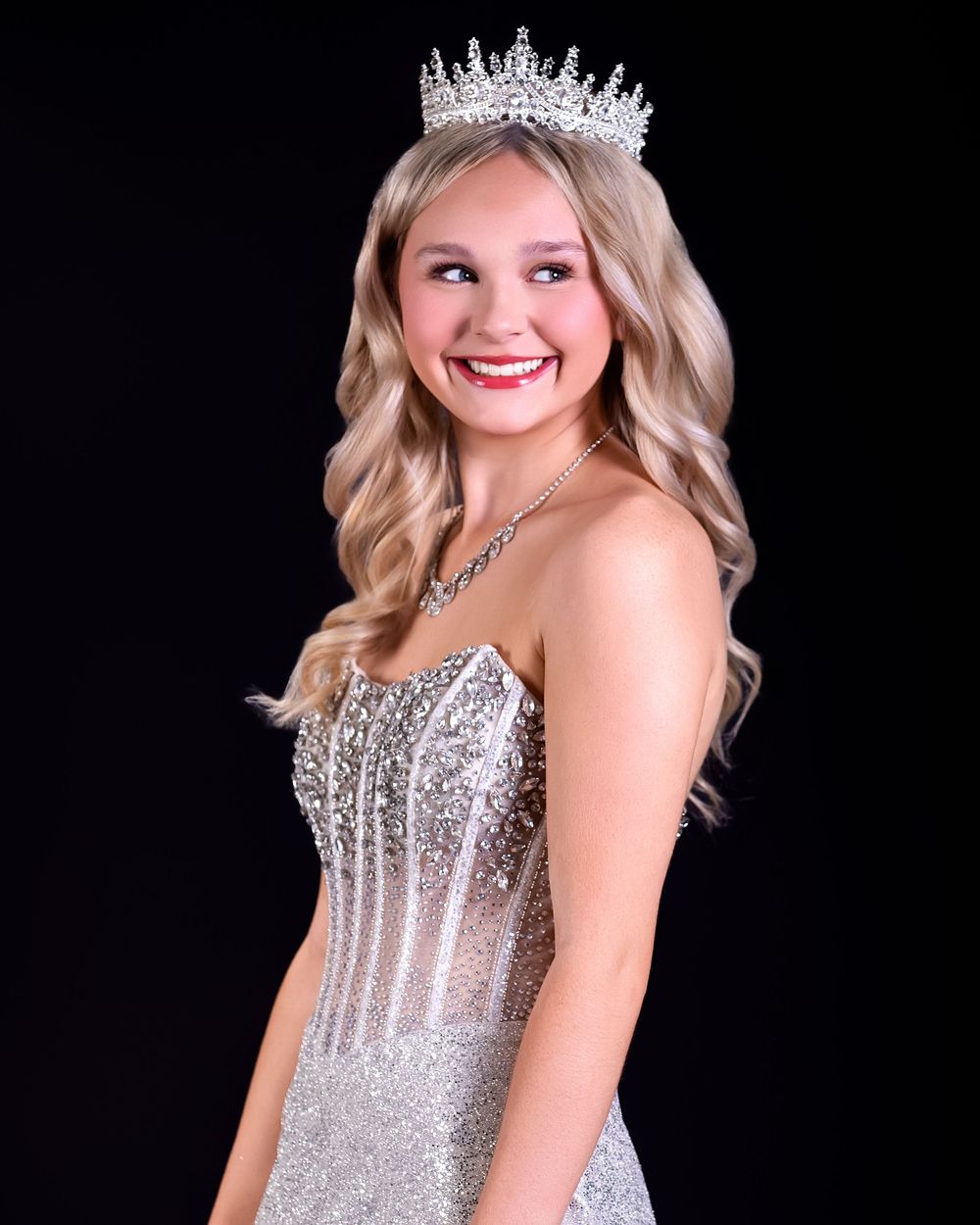 Young woman in sparkling silver gown and rhinestone crown smiling against black background.
