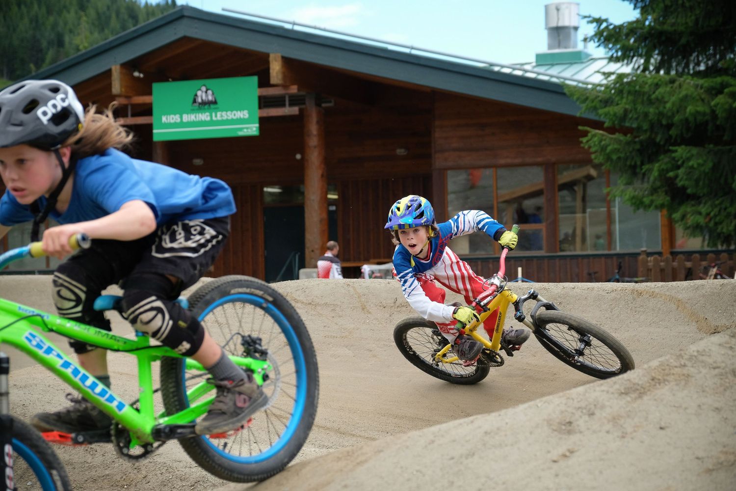 Young riders navigate berms on dirt track near wooden cabin at mountain bike park.
Whistler Mountain Bike Park