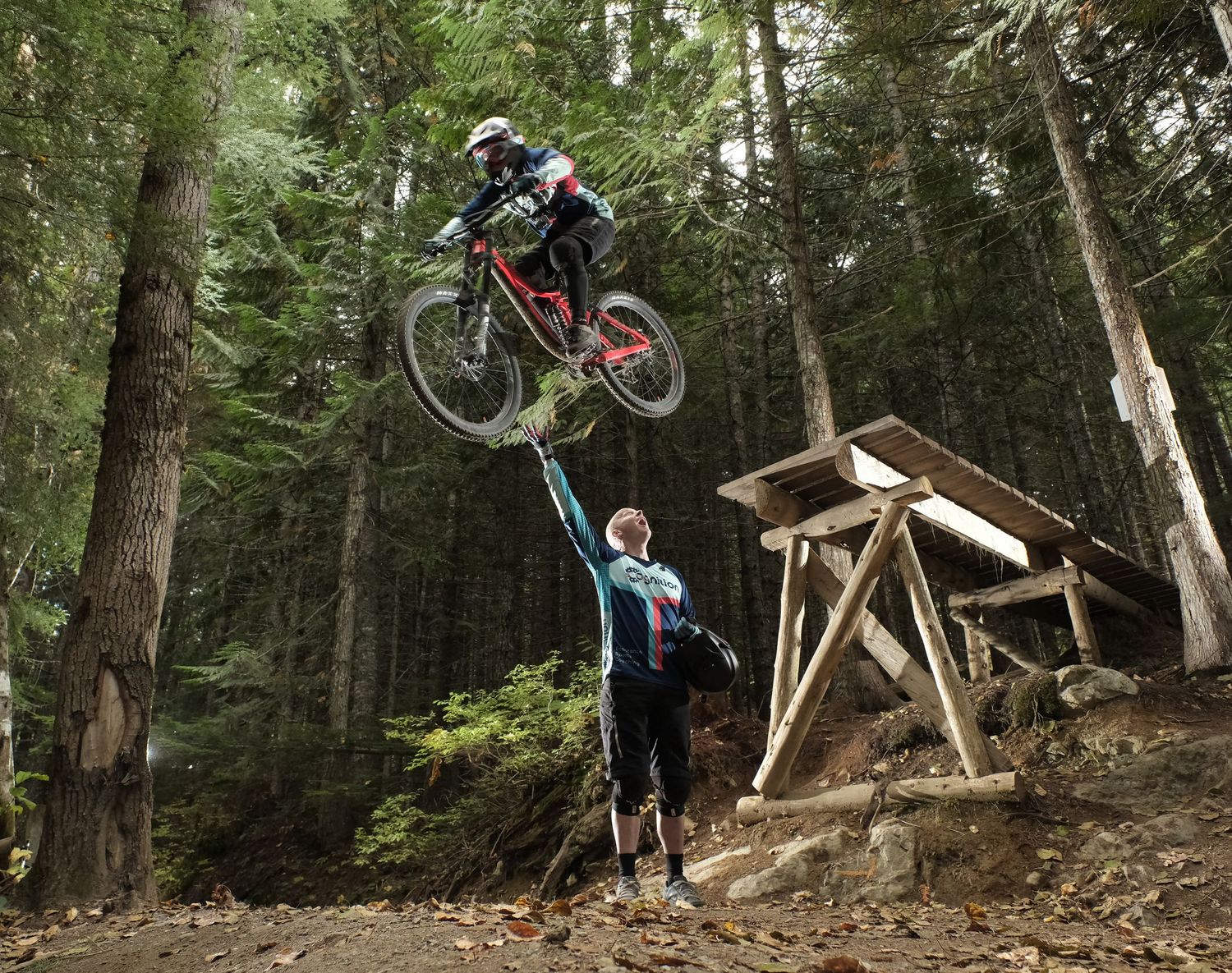 Mountain bikers perform aerial stunt over wooden ramp in dense forest.
Whistler Mountain Bike Park