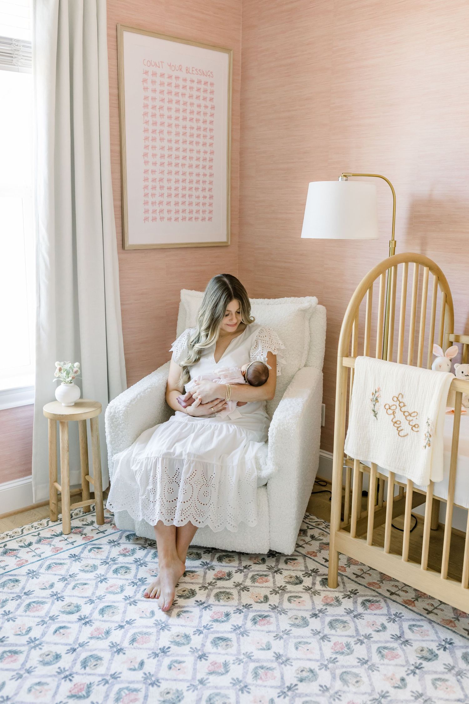 Peaceful nursery scene with pink walls, white chair, and wooden crib next to a table lamp.