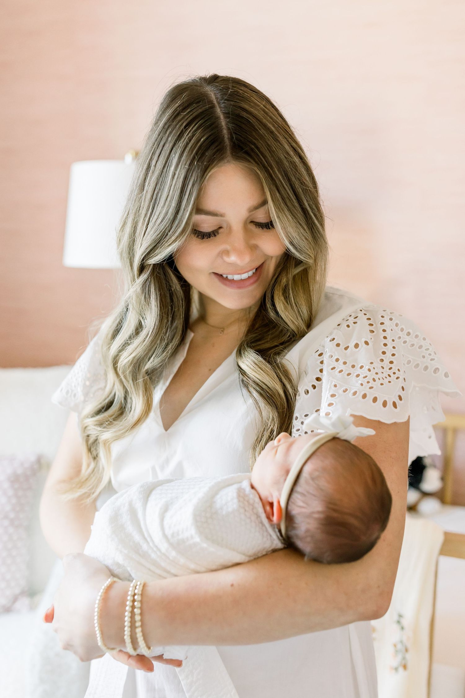 Mother in white eyelet dress holding sleeping newborn baby in nursery setting.
