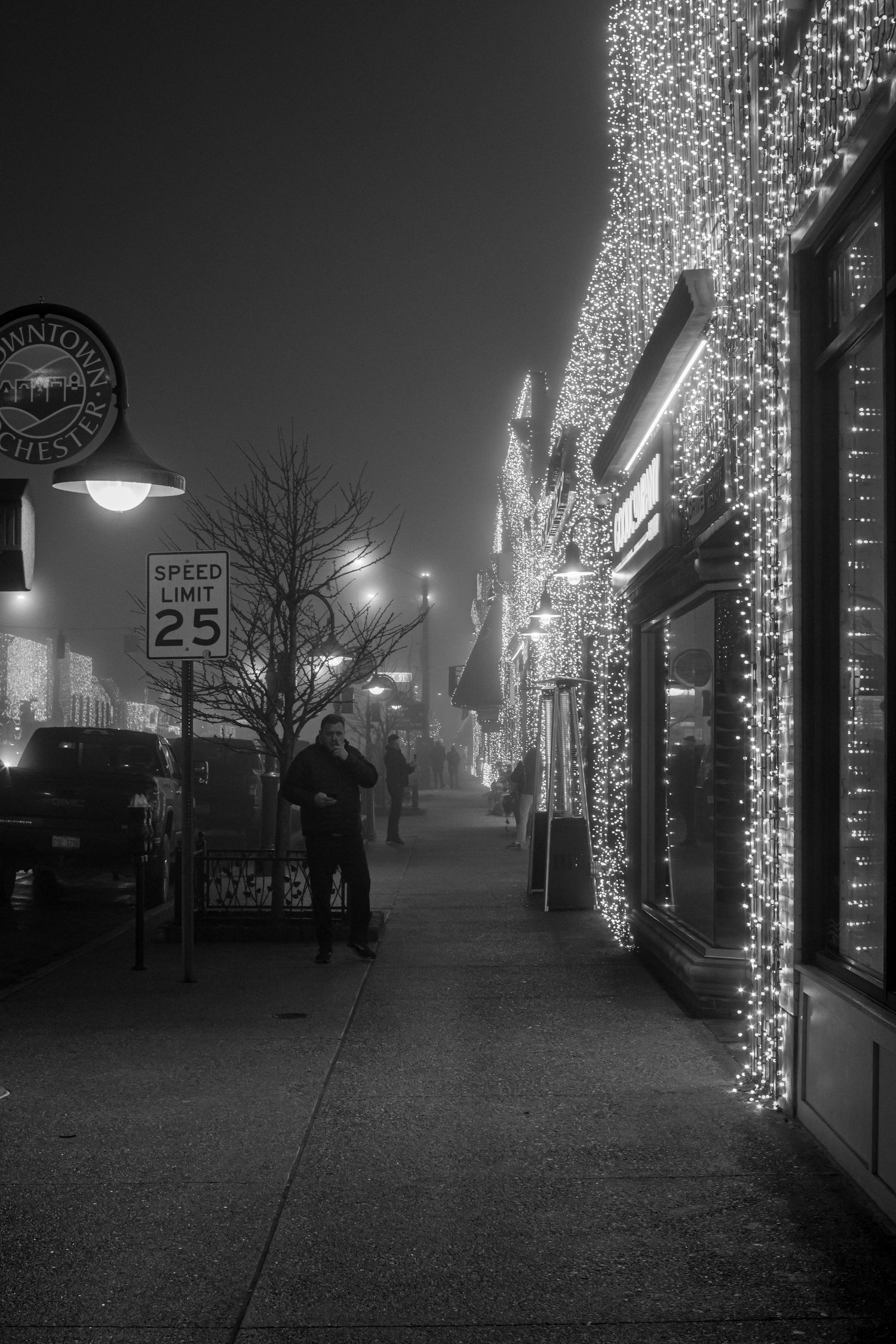 Downtown sidewalk with festive holiday lights and storefronts glowing on a misty evening.