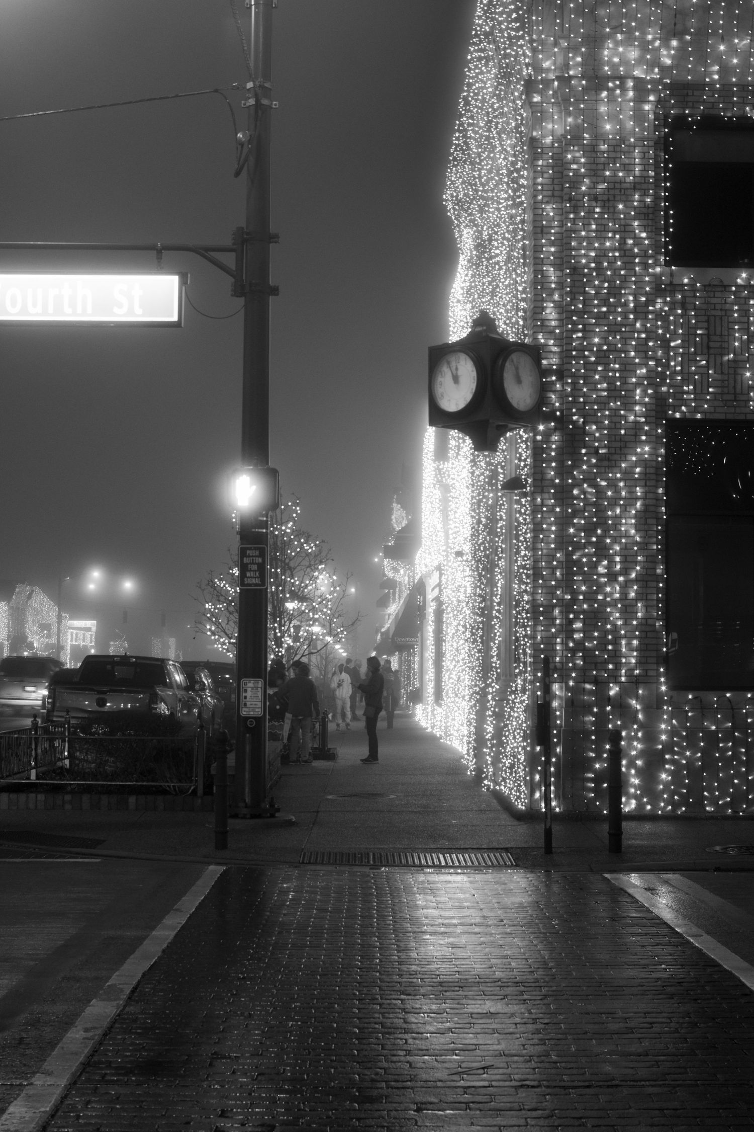 Street decorated with hanging lights and illuminated clock face visible through misty evening atmosphere.