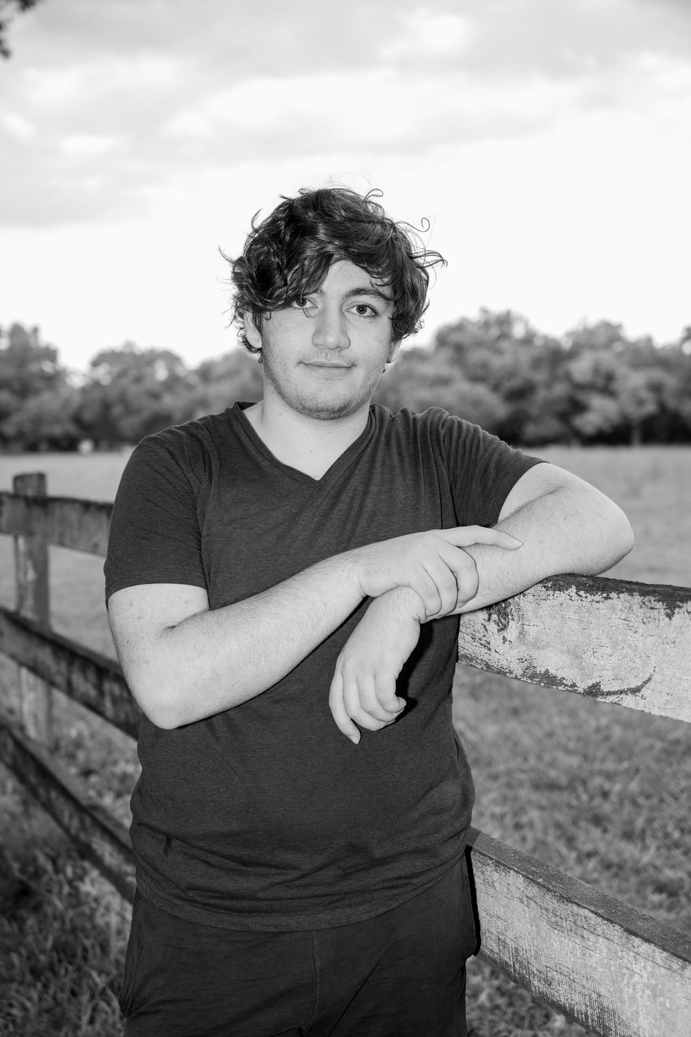 Black and white portrait of a young man with curly hair leaning on a wooden fence in a rural field.