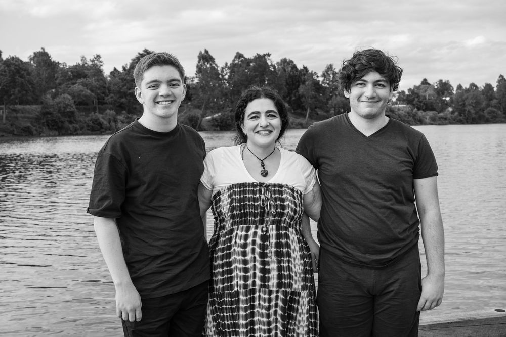 Black and white photo of three people smiling together near a lake with trees in the background.