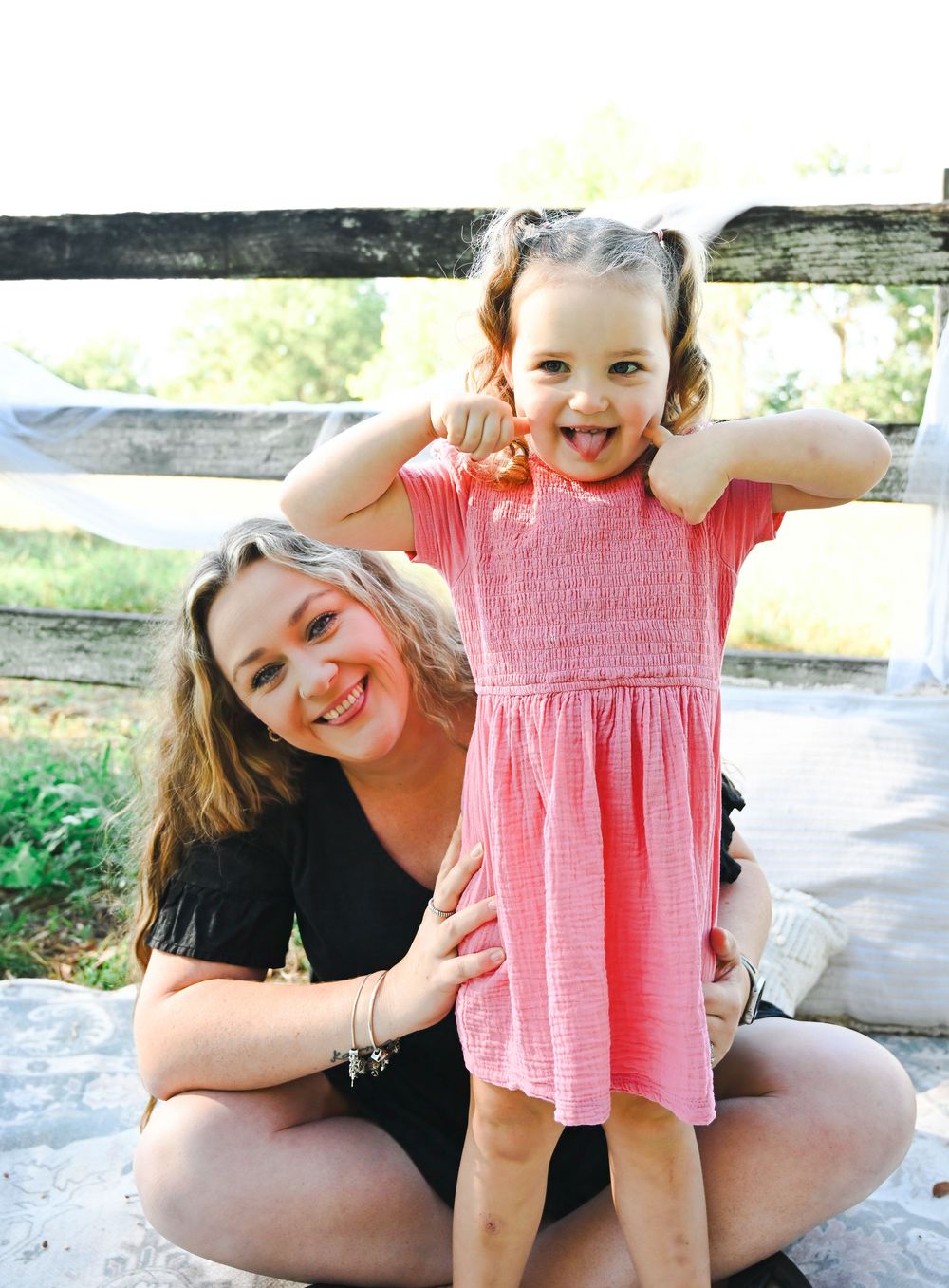 A smiling woman sits cross-legged outdoors while a happy young girl in a pink dress flexes her arms beside her.