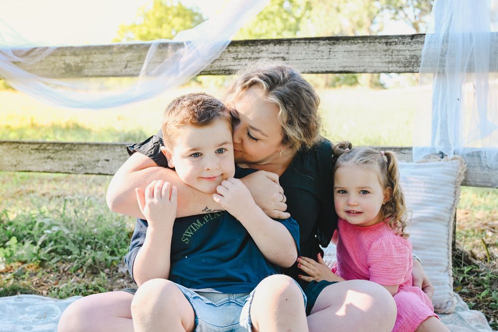 A mother lovingly kisses her young son while her toddler daughter sits beside them outdoors.