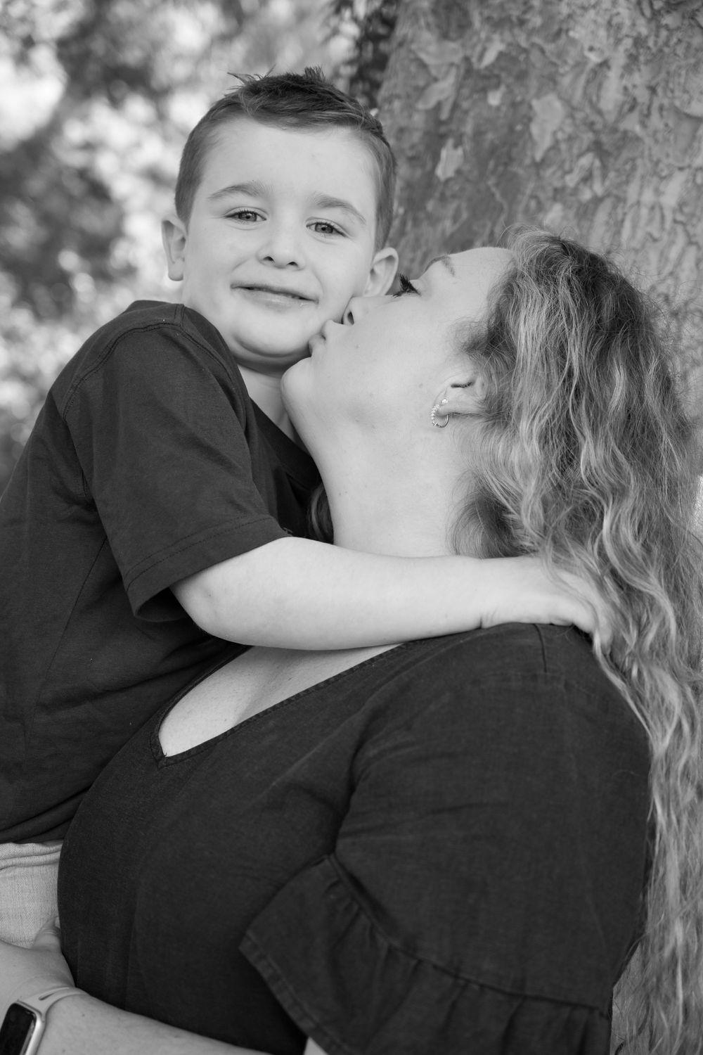 Black and white photo of a woman kissing a smiling young boy on the cheek outdoors near a tree.