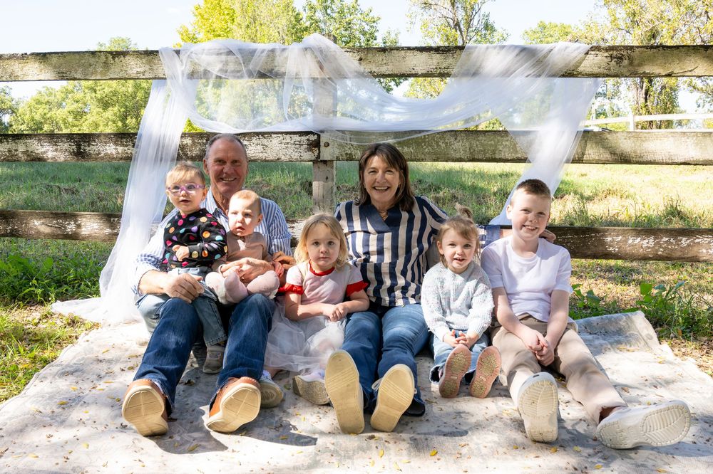 Family of seven posing outdoors on a blanket by a wooden fence, surrounded by lush green trees on a sunny day.