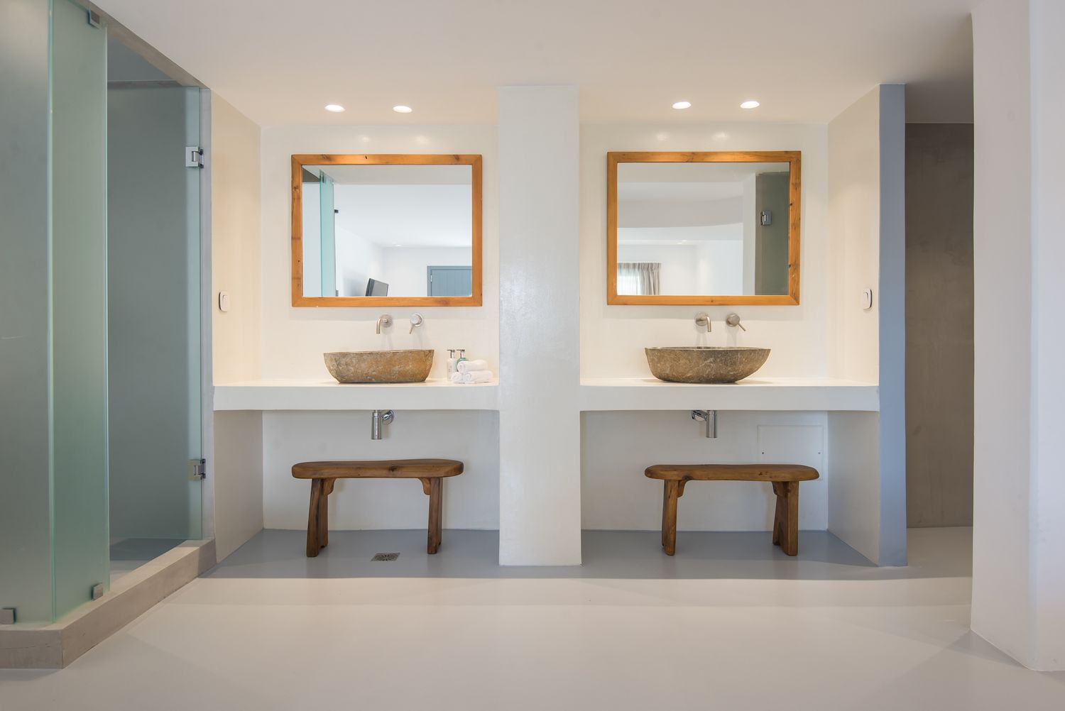 Modern minimalist bathroom with twin vessel sinks, wooden benches, and white walls creating a spa-like atmosphere.
