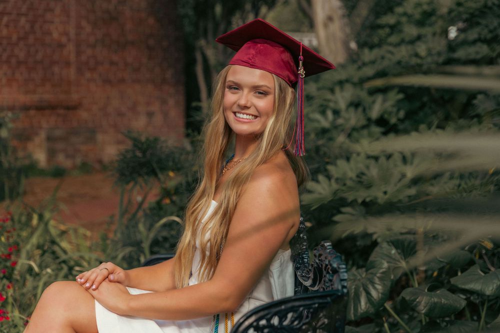 Graduate in red cap and white dress poses outdoors on decorative black bench with greenery in background.