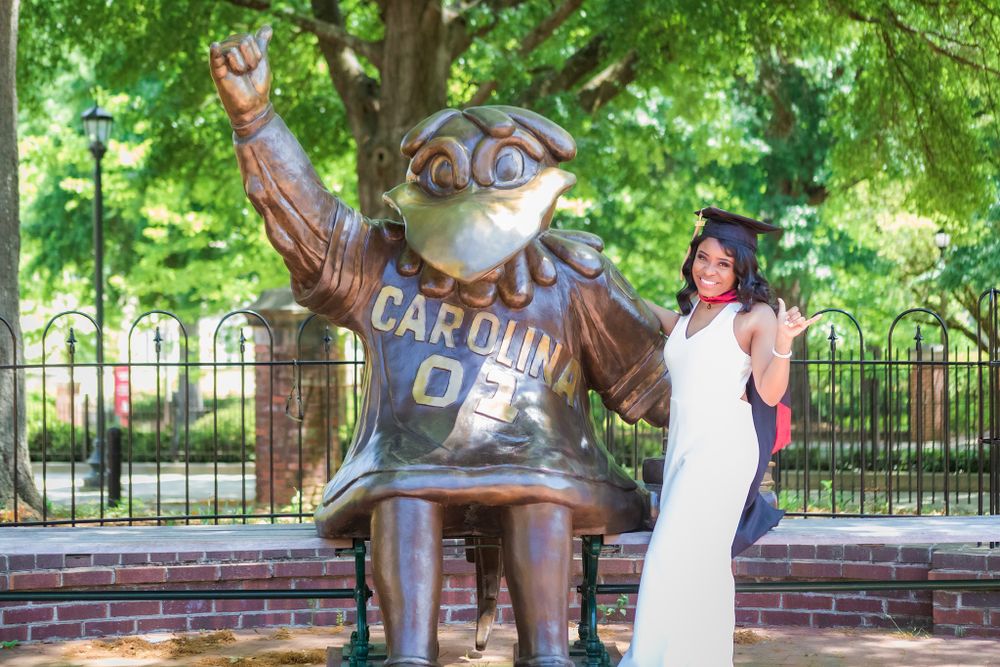 A person in white dress poses with Rameses mascot statue at UNC Chapel Hill campus on a sunny day.