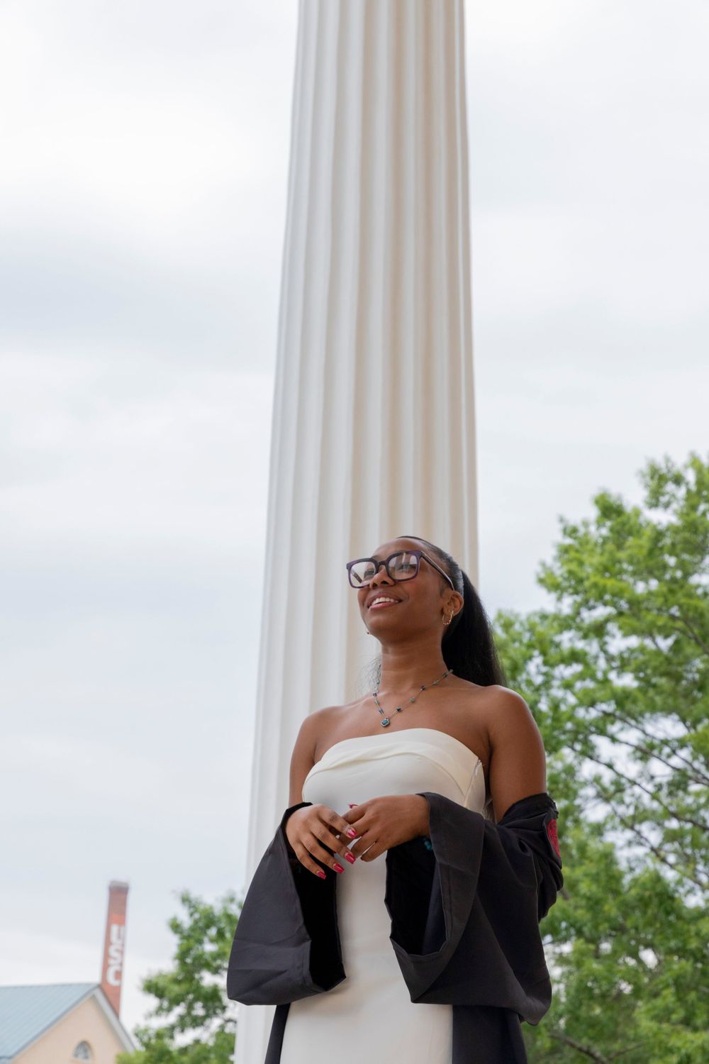 Low angle view of person in white dress and cardigan standing by classical white column against cloudy sky.