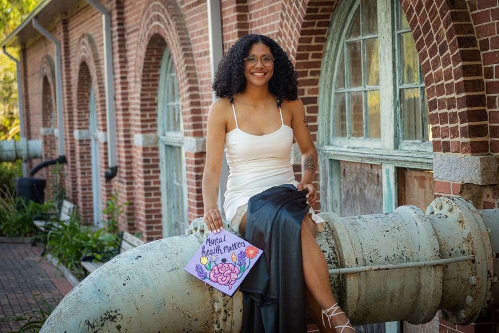 Someone in a white top and dark skirt sits on a decorative cannon outside a historic brick building with arched windows.