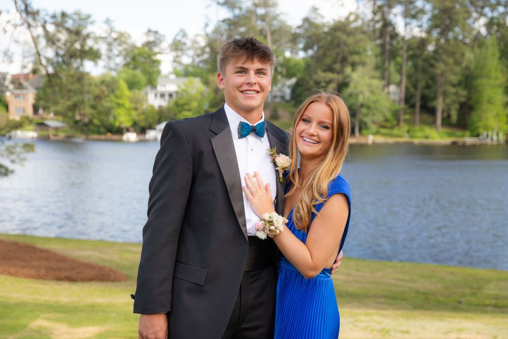 A couple in formal attire poses near a scenic lake, with the person wearing a black tuxedo and blue bow tie.