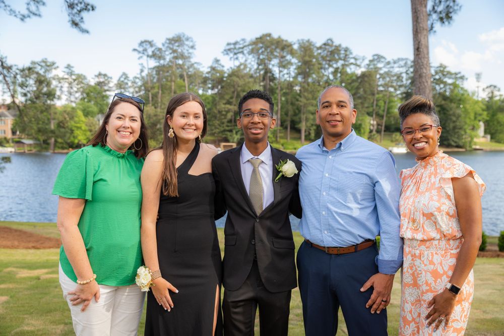 A group of five people stand together smiling by a scenic lake with trees in the background.