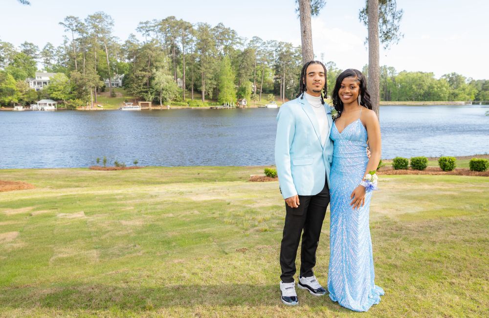 Two people in formal attire pose together on a grassy lawn beside a scenic lake with trees in the background.