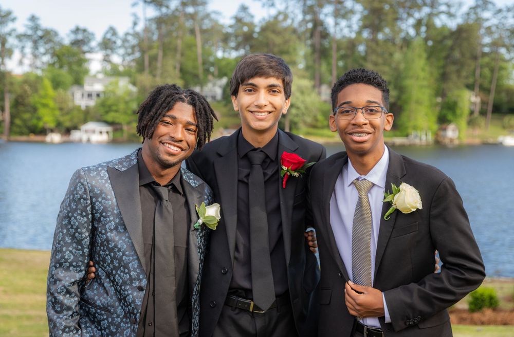 Three friends wearing formal attire pose together outdoors on a sunny day near a lake with trees in the background.