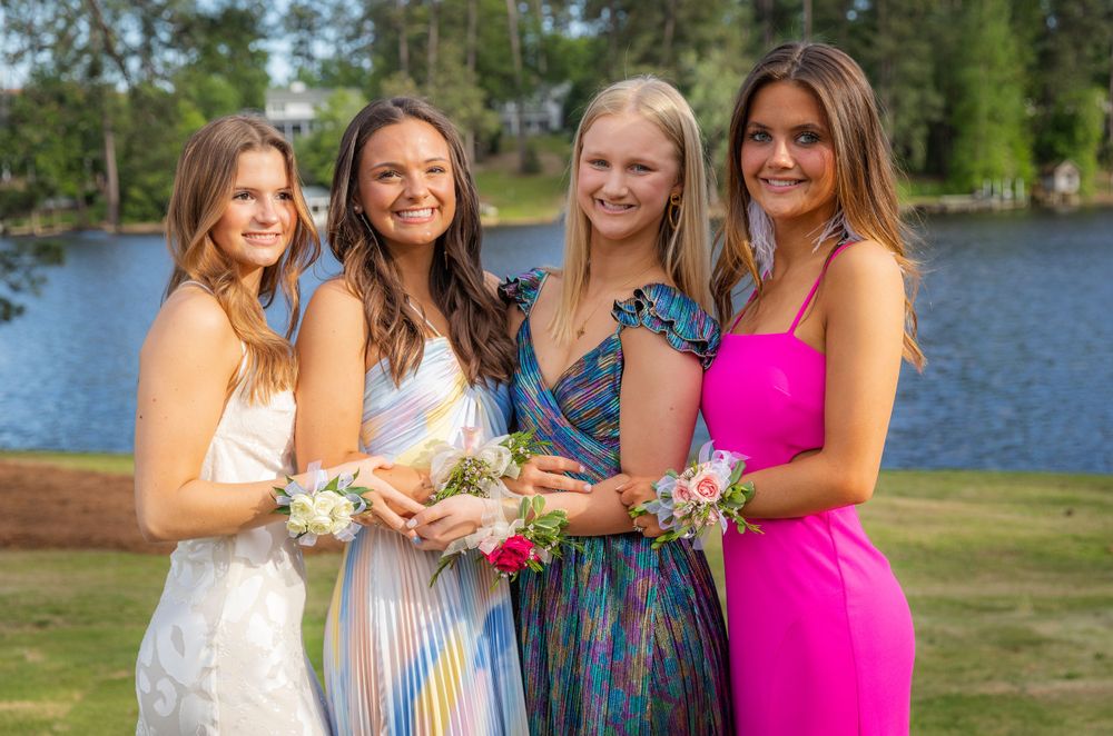 Four people in formal attire stand together smiling by a lake with trees and flowers on a sunny day.