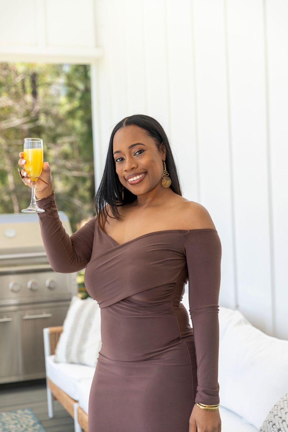 Woman in a brown off-shoulder dress holds up a glass of orange juice while smiling on an outdoor patio.