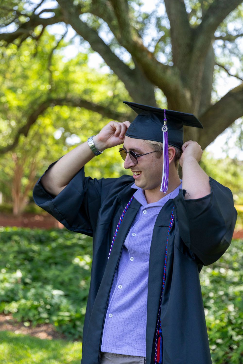 Graduate adjusts mortarboard cap during outdoor graduation photoshoot in sunny garden setting.
