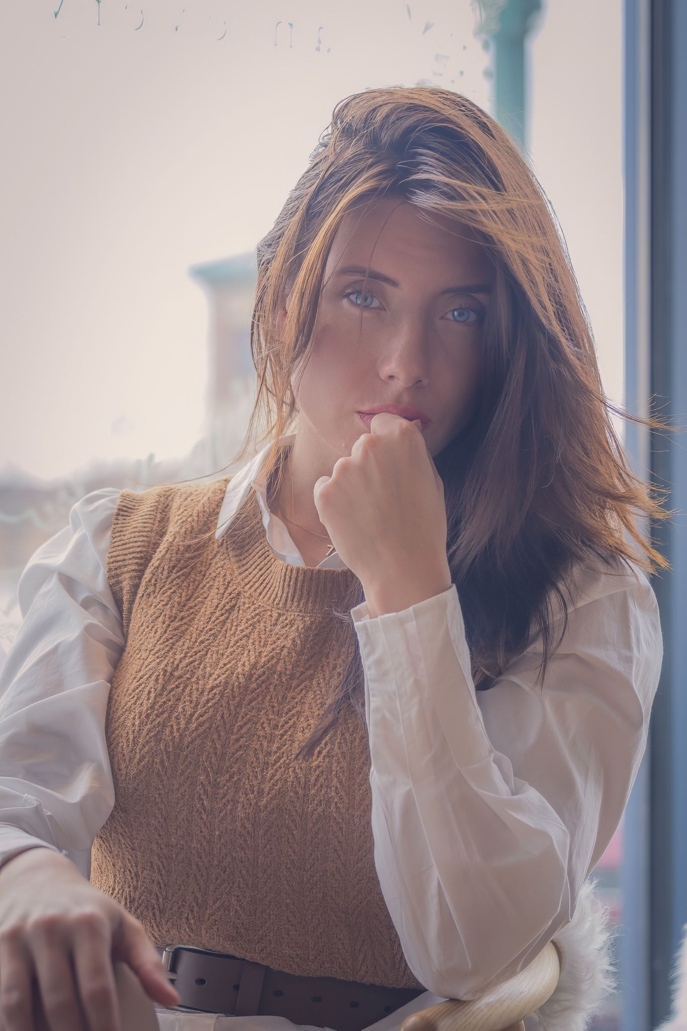 Thoughtful looking person wearing a brown knit vest and white blouse poses near window in soft lighting.