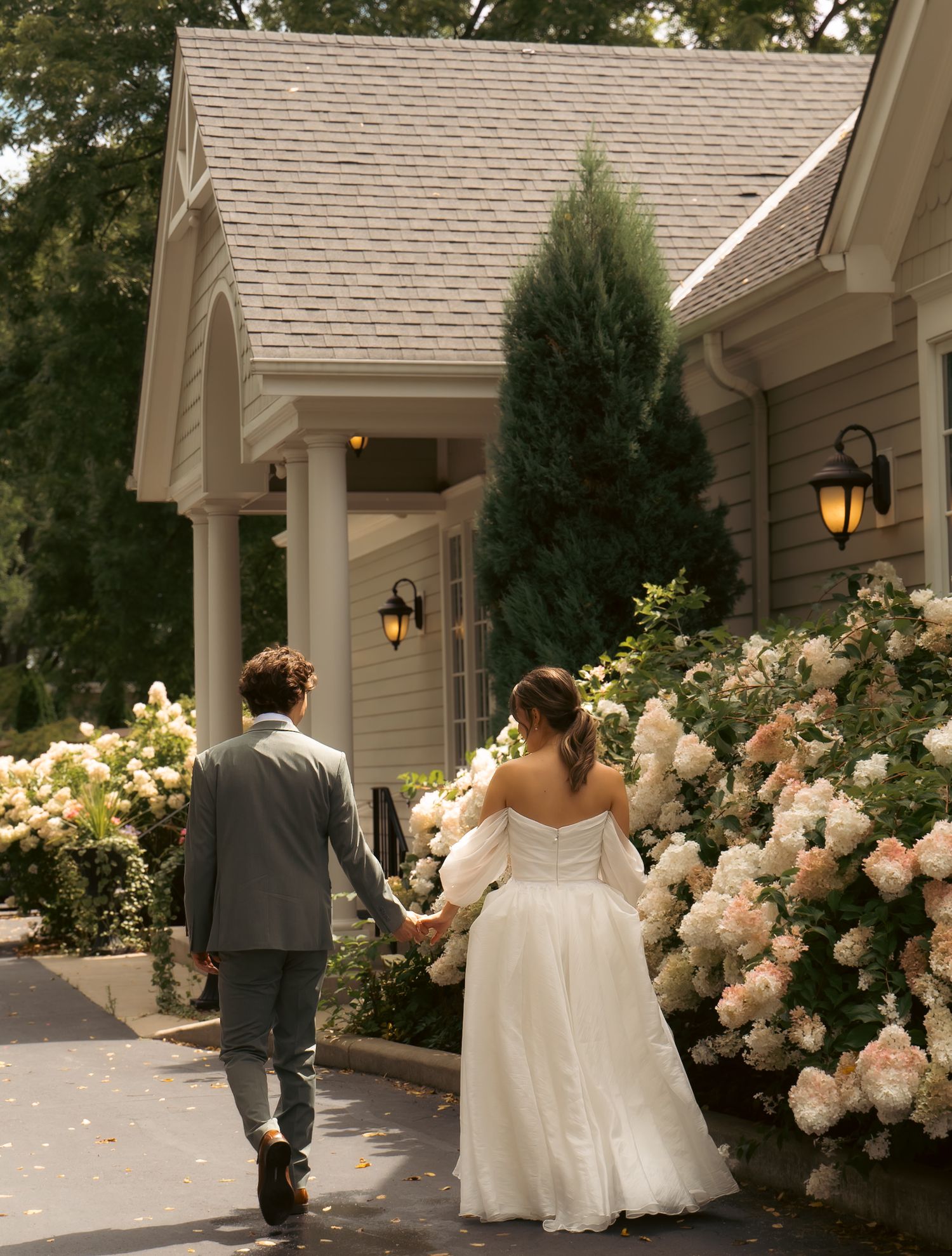 A couple walks hand in hand past blooming roses toward a house entrance with lanterns.