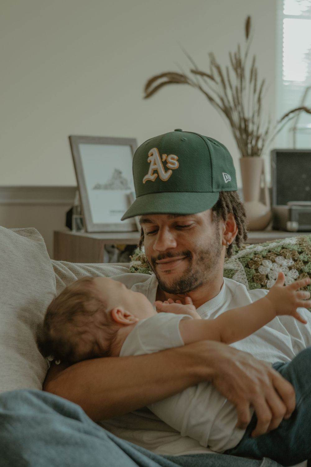 A tender moment of affection between a parent wearing an Oakland A's cap and young child on a couch.