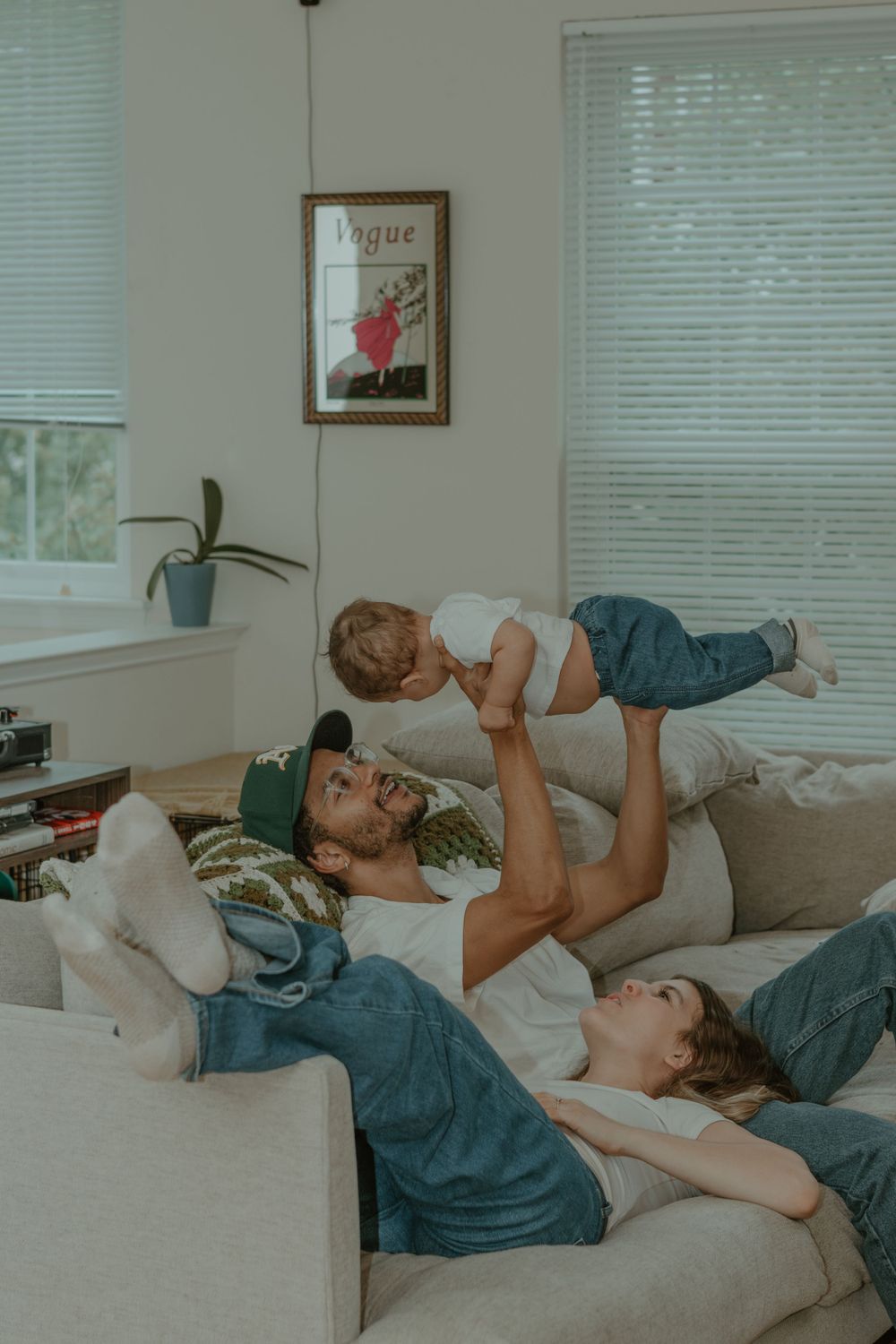 Playful bonding moment as parent lifts child in air while relaxing on couch with Vogue poster in background.