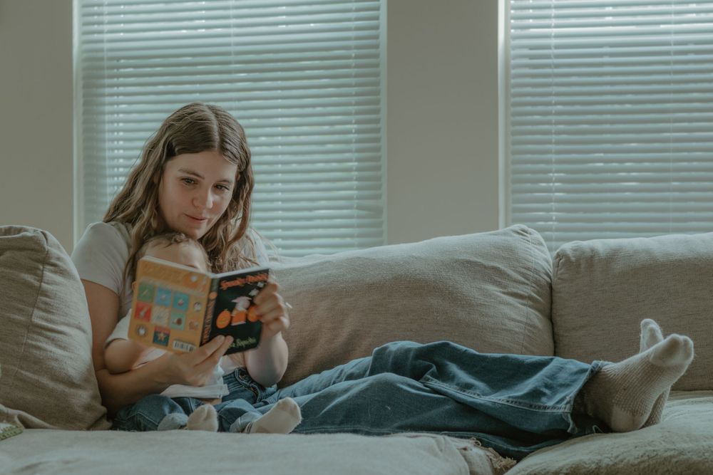 Person relaxing on gray couch while reading a book in afternoon light.