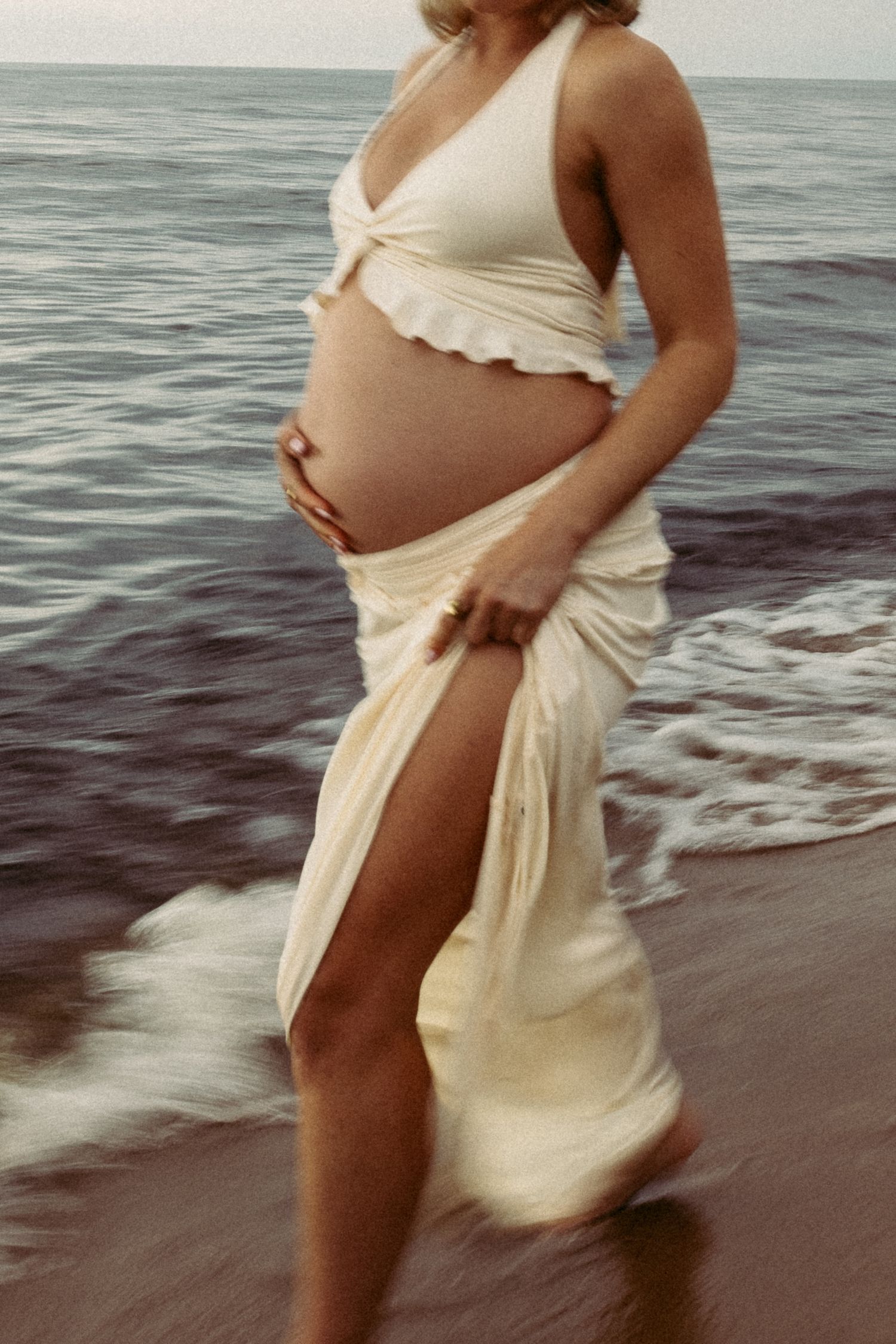 Woman in white dress walking along beach shoreline at sunset.