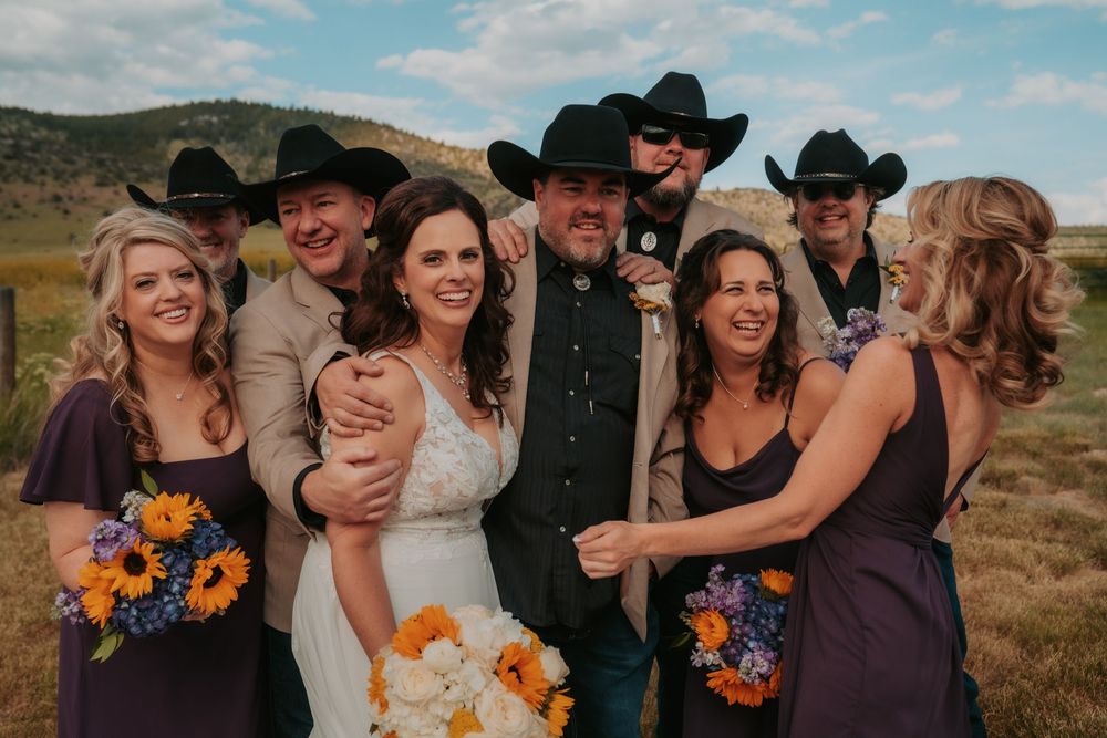 A wedding party with bridesmaids in purple dresses and groomsmen in cowboy hats pose together against a mountain backdrop.