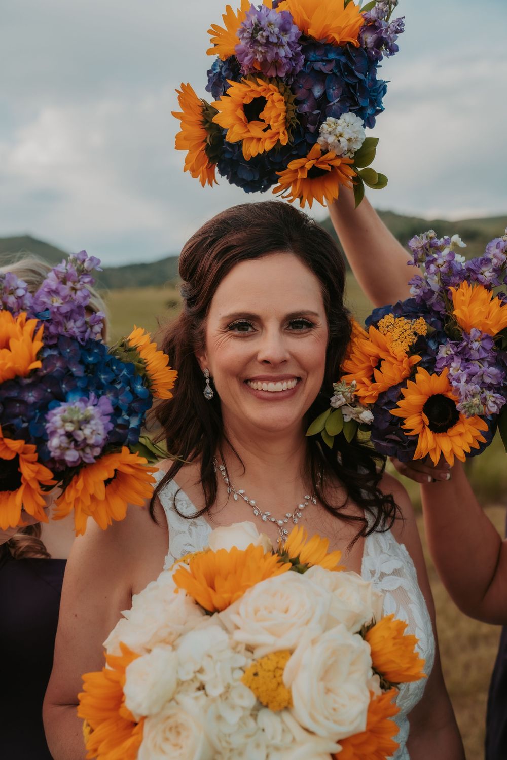 A vibrant bridal bouquet features sunflowers, purple flowers and white roses against a rolling hills landscape.
