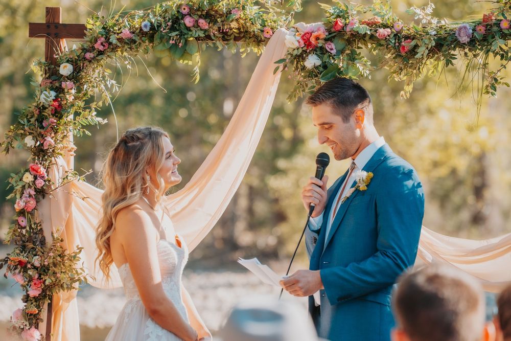 A romantic outdoor wedding ceremony under a floral arch decorated with pink and red roses and flowing fabric.