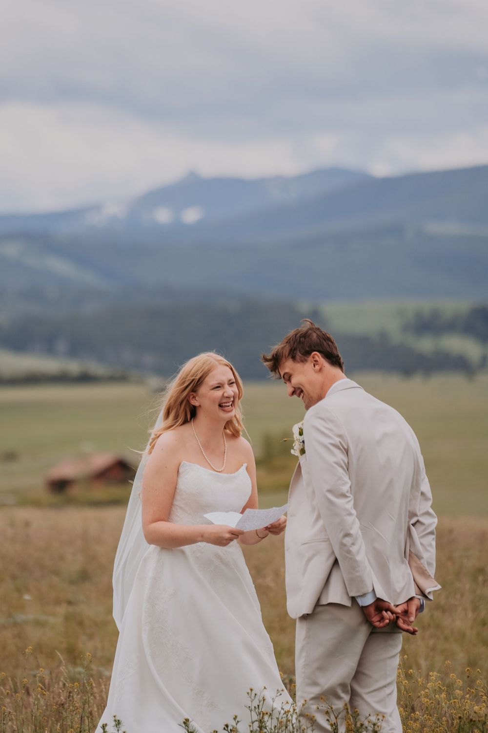 A bride and groom share a joyful moment reading vows in a scenic mountain meadow on their wedding day.
