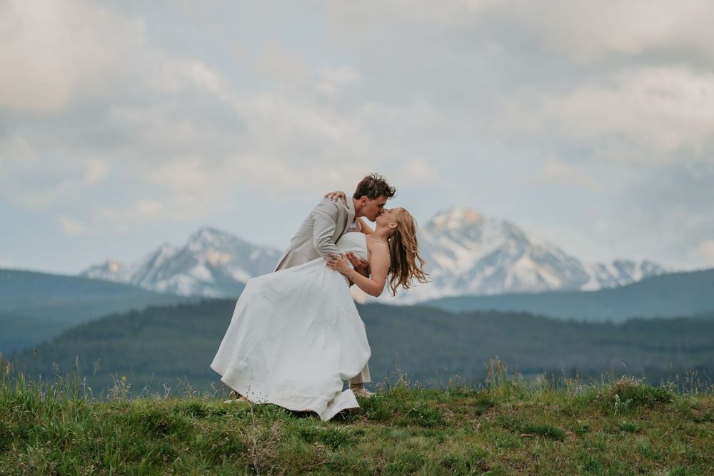 Bride and groom share a romantic dip kiss on a mountain meadow with snow-capped peaks in the background.