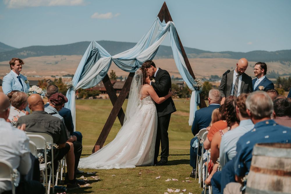 A rustic outdoor wedding ceremony takes place under a wooden arch draped with flowy fabric against rolling hills and mountains.