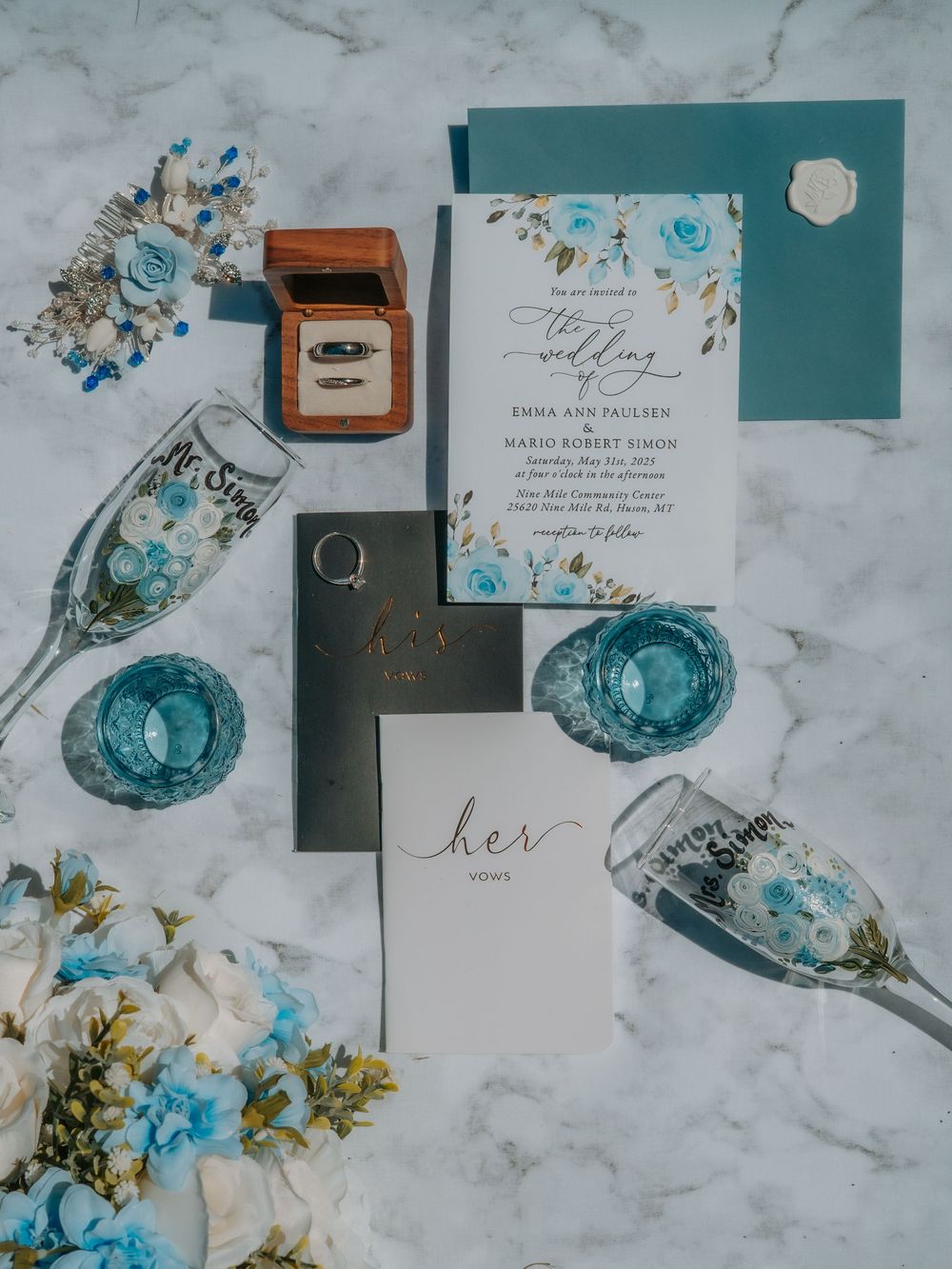 Wedding invitation suite with teal and blue floral design alongside rings, glasses and decor on marble surface.