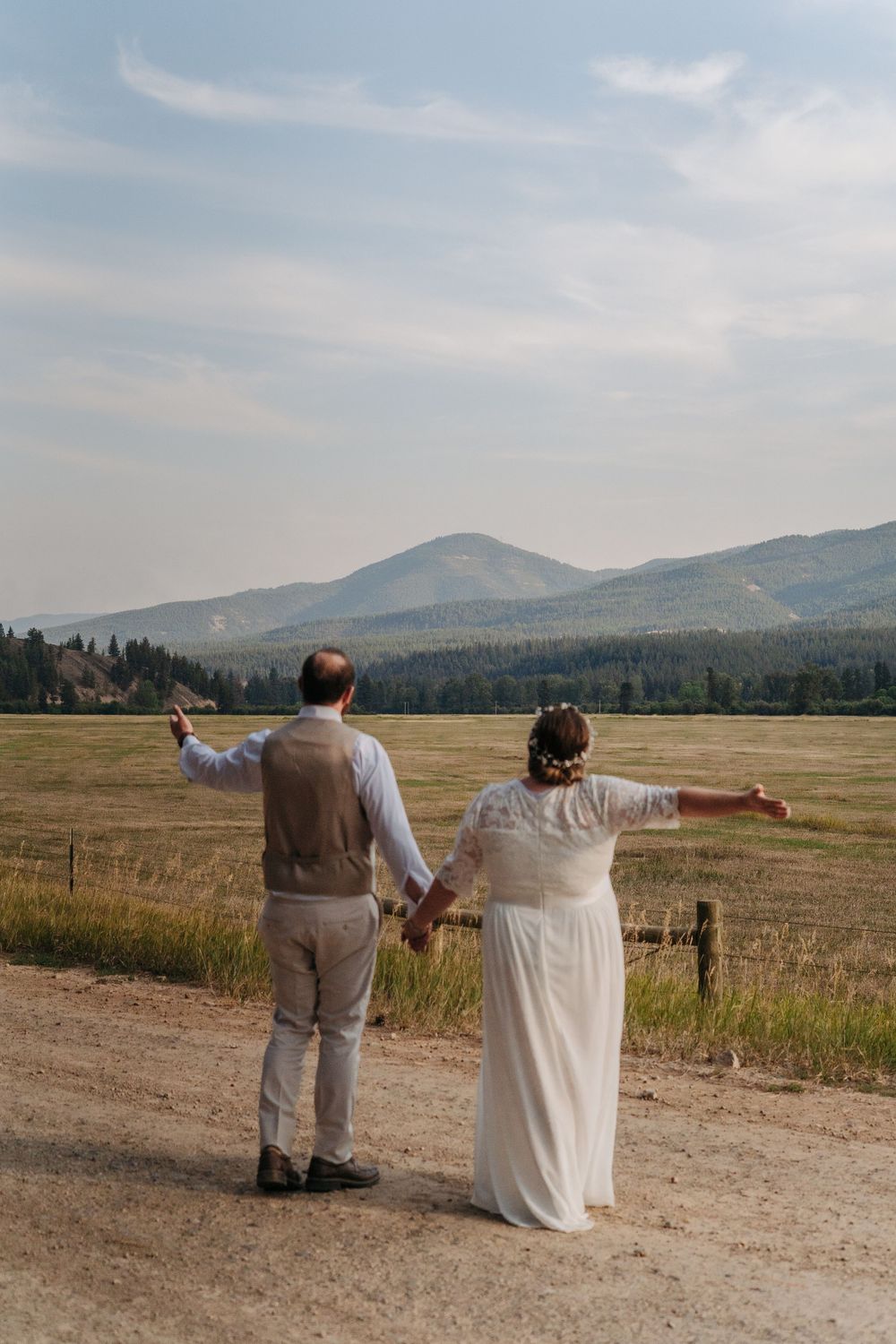 A couple in wedding attire stand on a dirt road overlooking scenic mountain views in the distance.