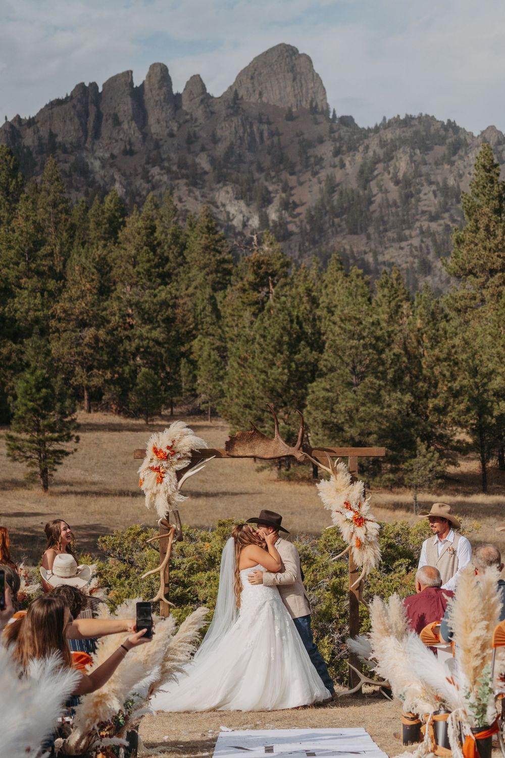 A wedding celebration takes place in a scenic mountain landscape with evergreen trees and rocky peaks in the background.