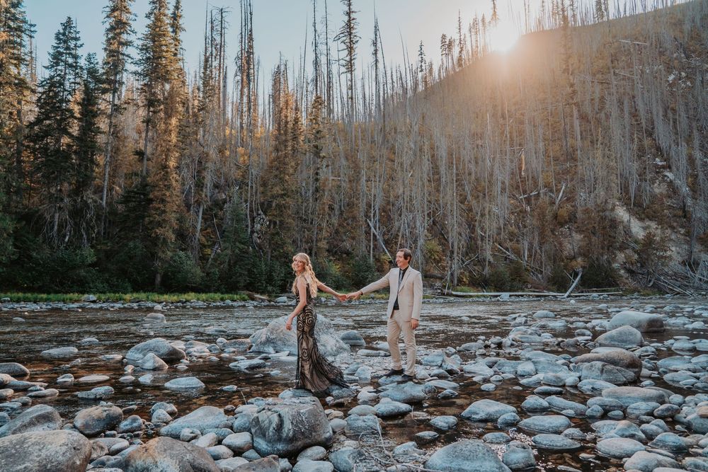 A couple walks hand in hand through a snowy winter forest landscape with sunlight streaming through bare trees.