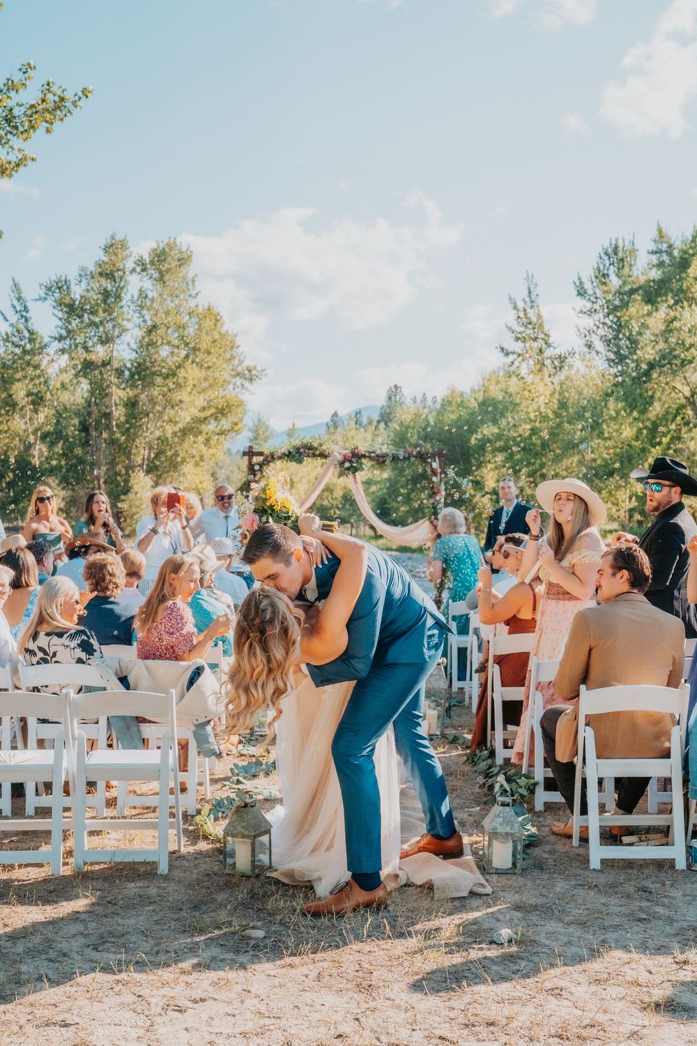 A joyous outdoor wedding ceremony with guests sitting in white chairs as a couple shares a romantic dip kiss in golden sunlight.