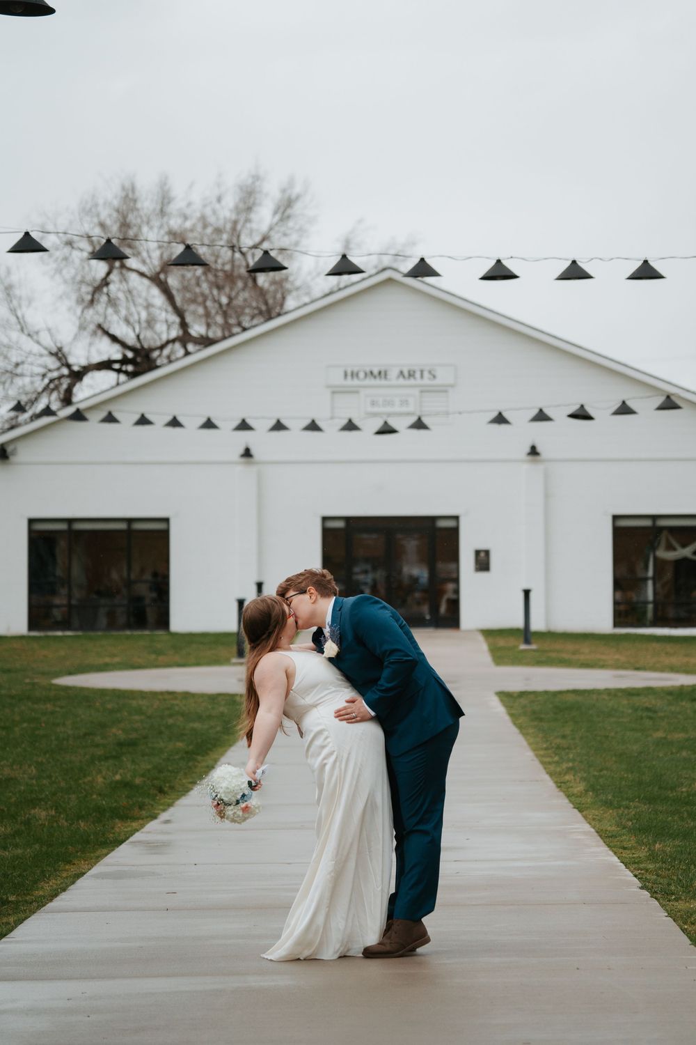 A couple shares a romantic dip kiss in front of a white building with "Home Arts" signage on a cloudy day.