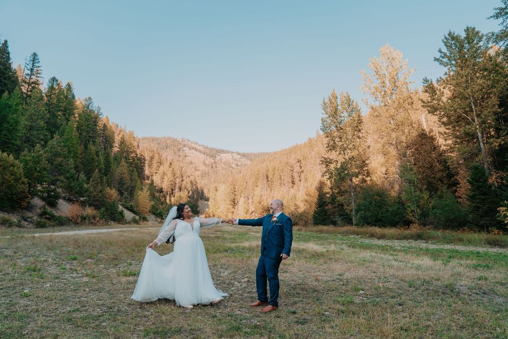 Couple in wedding attire stands in a scenic mountain meadow during golden hour with evergreen trees in the background.