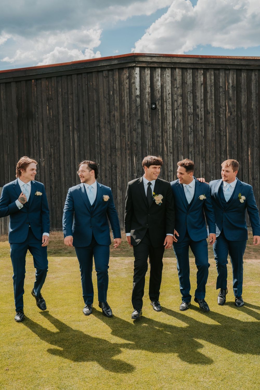 A group of groomsmen in navy and black suits walking and laughing together on grass in front of a rustic barn.