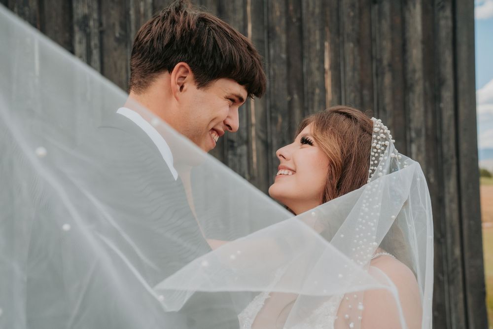 A joyful couple shares a loving moment under a flowing white veil against a rustic wooden wall backdrop.