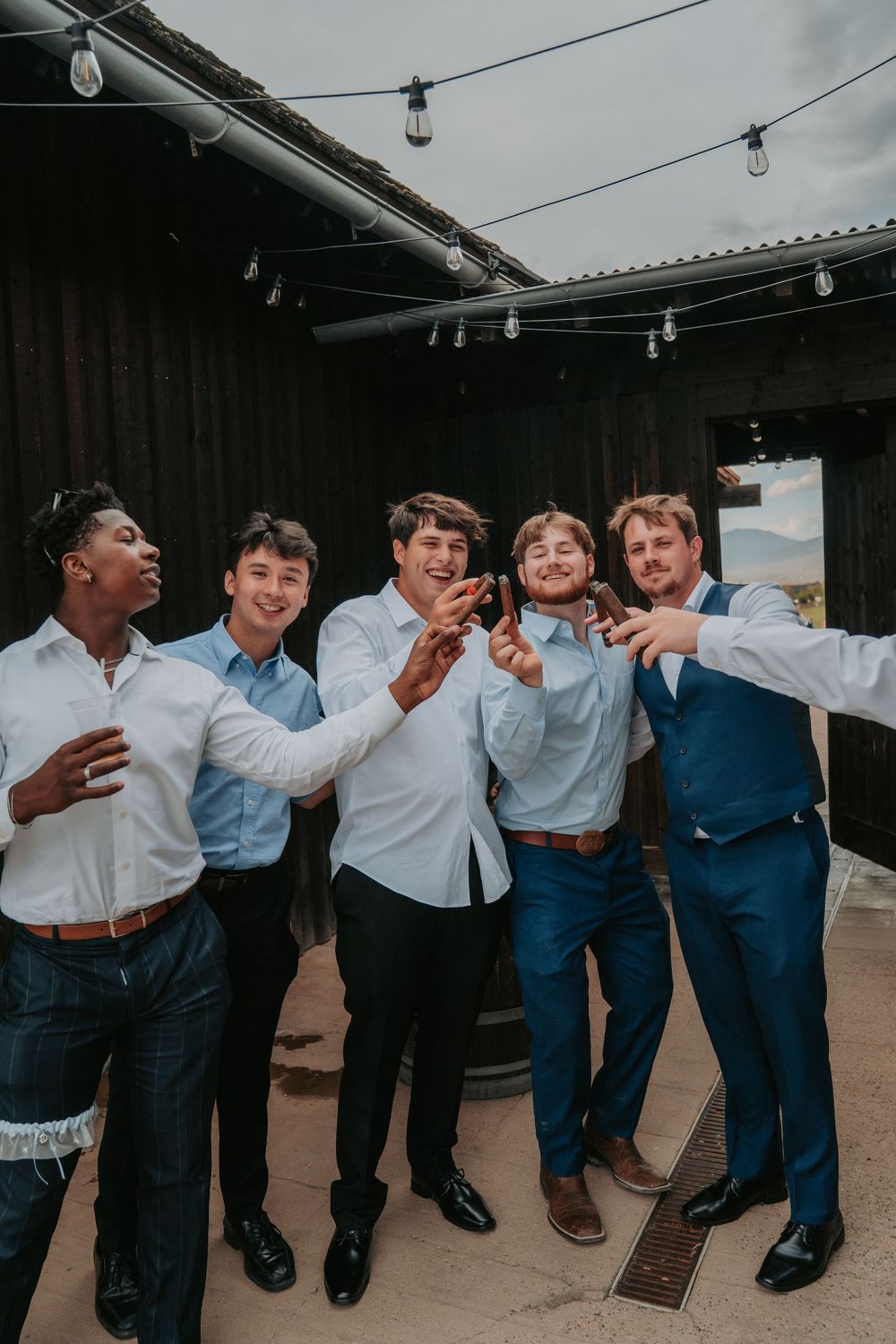 A group of friends in formal attire laugh and celebrate together under string lights at an outdoor venue.