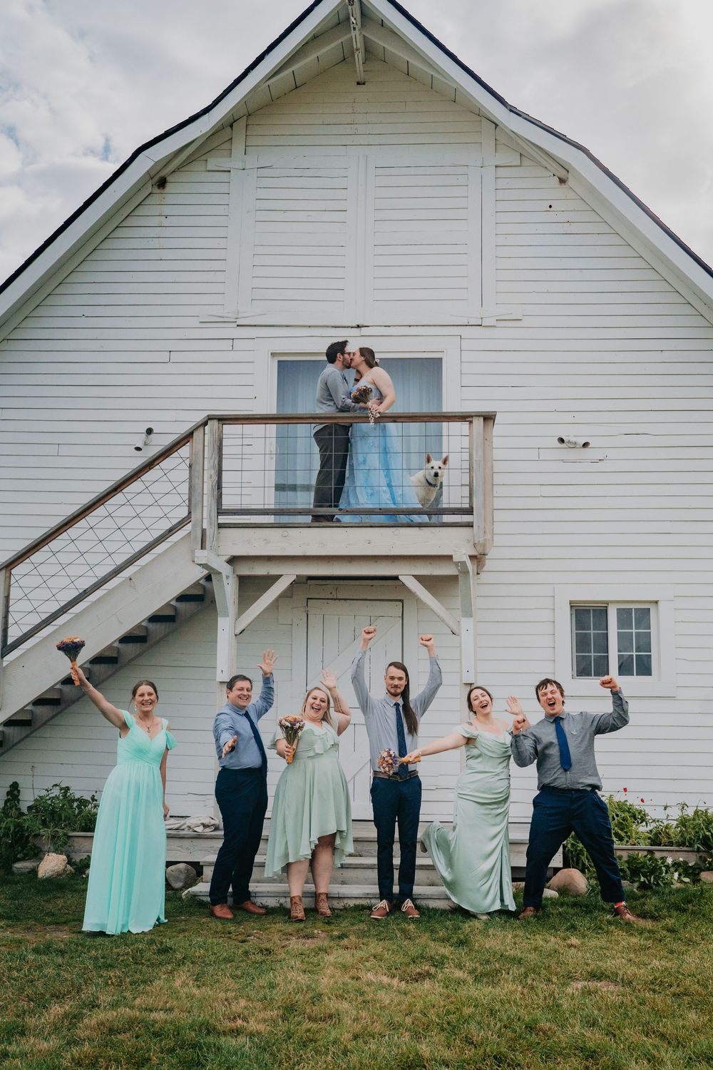 A fun wedding party poses in a sequence of playful jumping shots against a white barn backdrop.