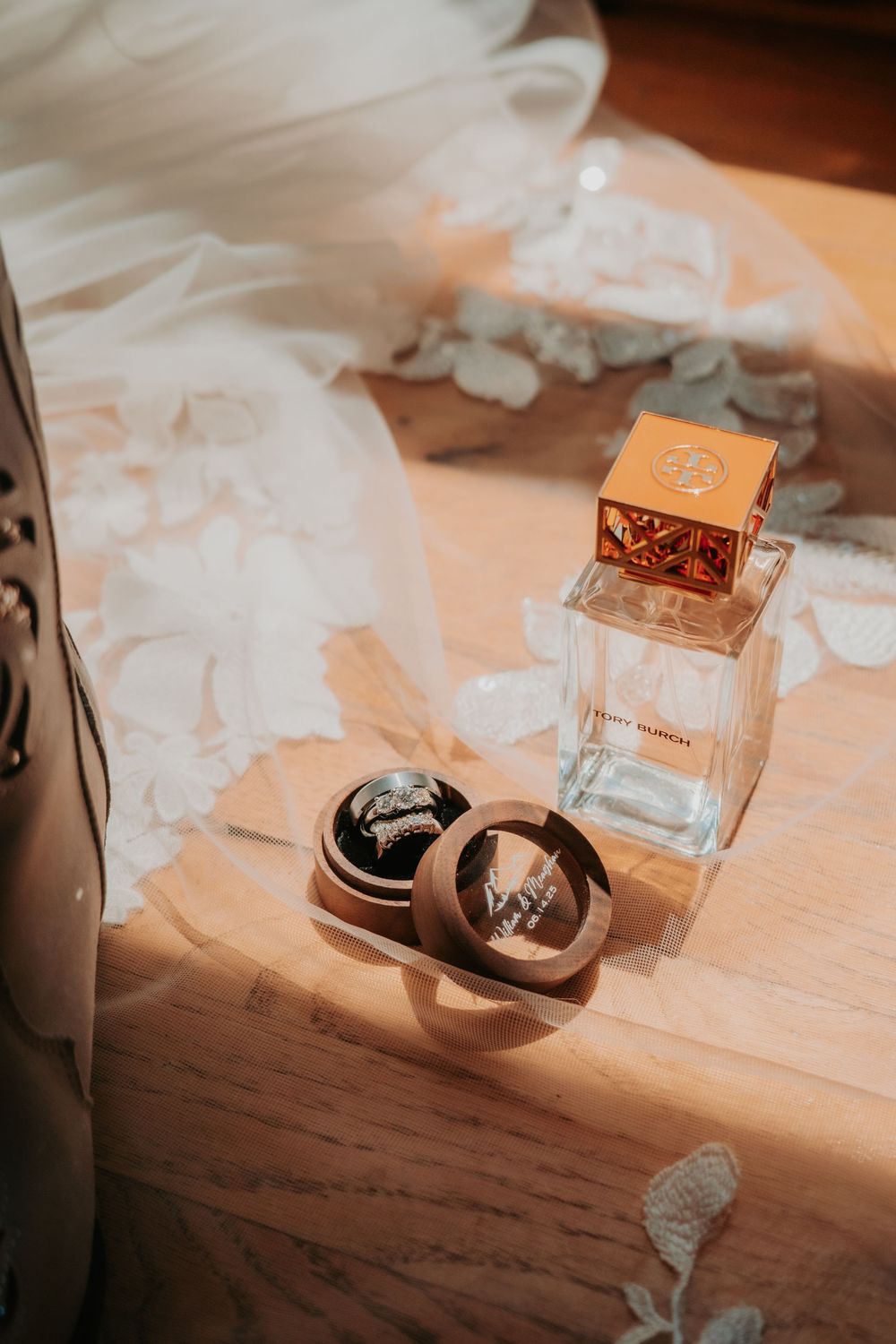 Wedding rings in a small box next to a perfume bottle with white tulle fabric and sunlit wooden surface.